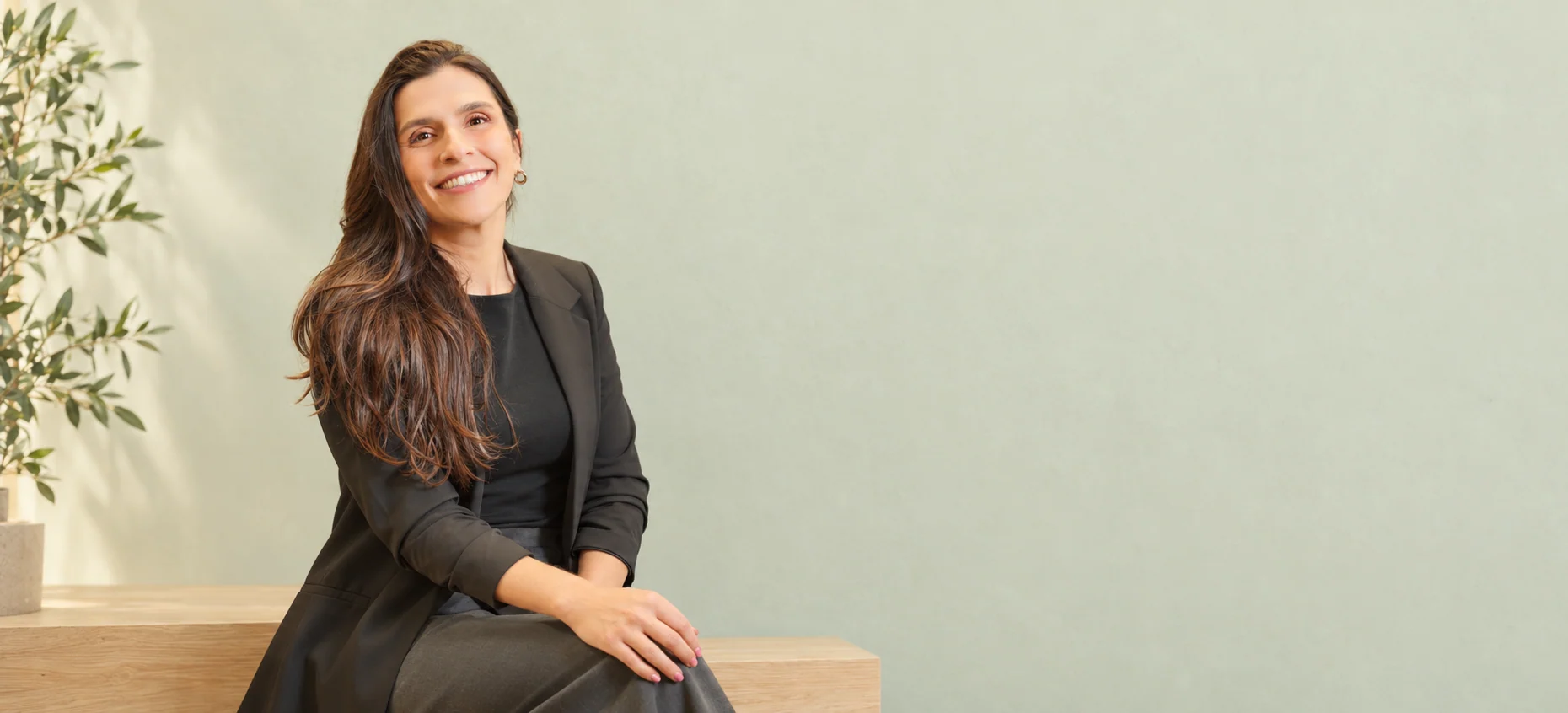 Dra. Gabriela Chefaly sorrindo, sentada de blazer preto em um cenário sálvia com luz natural.